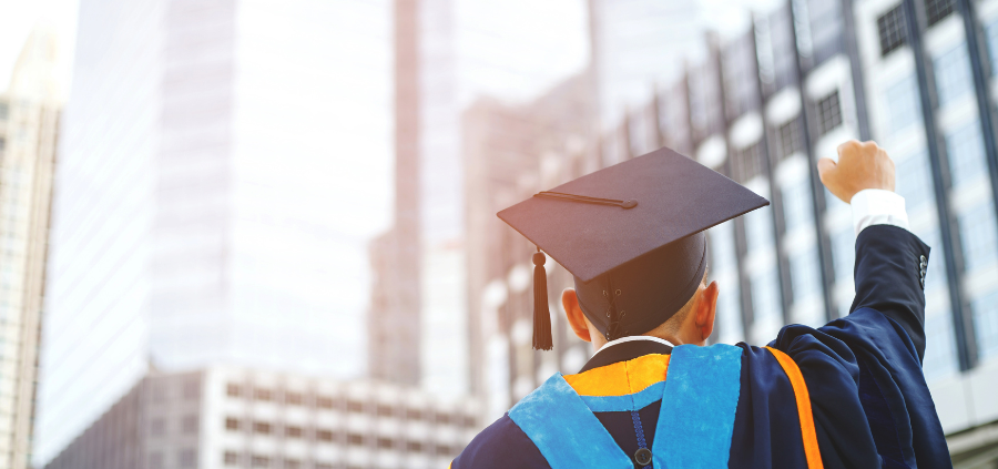 Man in graduation cap raises fist toward tall buildings