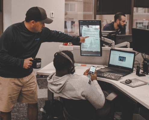 Two men working at a desk while one points at a computer and one sits down in the chair