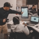 Two men working at a desk while one points at a computer and one sits down in the chair